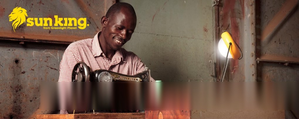 A tailor smiles as he works at his sewing machine beside a Sun King solar lamp